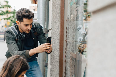 Young man photographing store window with mobile phone while standing with friend in city