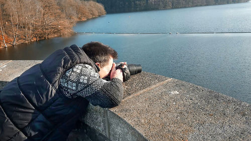 Side view of man photographing on retaining wall against river