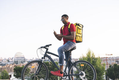 Delivery man wearing sunglasses text messaging through smart phone while standing with bicycle