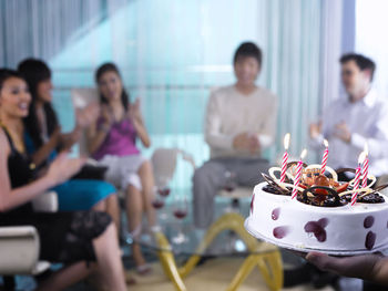 Cropped hand of person holding birthday cake against young friends enjoying party