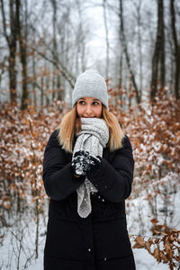 Portrait of young woman wearing warm clothes while standing in forest during winter