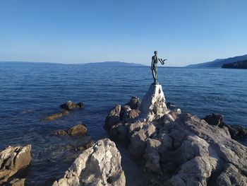 Statue on rock formation in sea against clear blue sky
