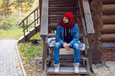 Man with a hipster beard in a red knitted hat and a denim jacket is sitting on the stairs