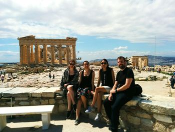 Portrait of happy friends sitting at parthenon against cloudy sky on sunny day