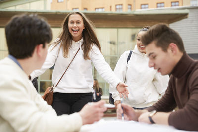 Happy teenage girls walking towards male friends sitting at table in schoolyard