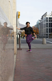 Woman listening music while exercising on sidewalk against sky in city