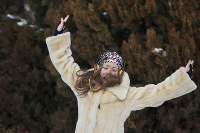 Cheerful young woman with arms outstretched listening music while standing against trees in park during winter