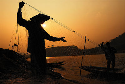 Silhouette men holding fishing net against sky during sunset