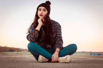 Portrait of young woman sitting at skateboard park against clear sky