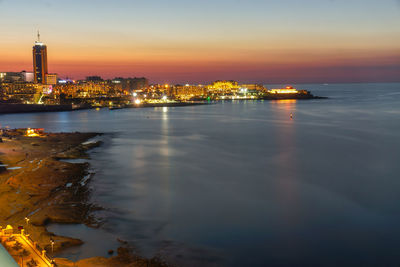 Illuminated city by river against sky at sunset