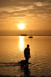 Silhouette man with dog standing in sea against sky during sunset