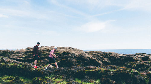 Side view of men walking on rocks against cloudy sky at looe during sunny day