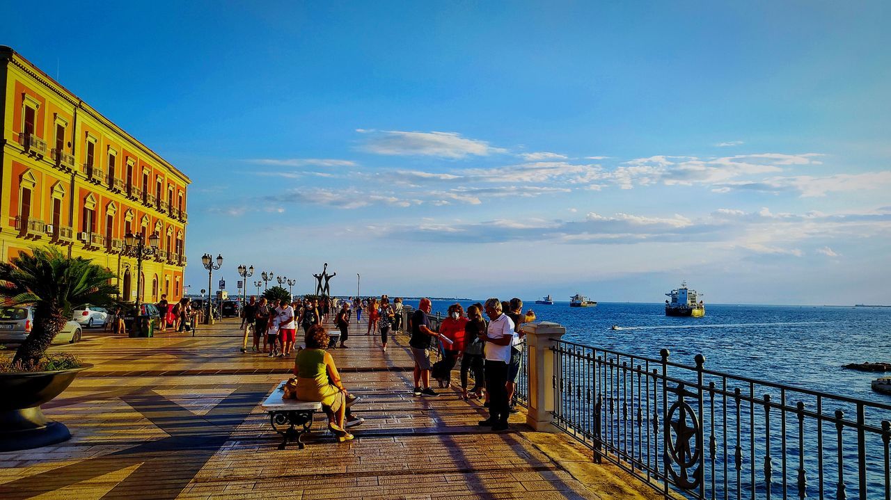 sky, water, vacation, architecture, walkway, sea, group of people, boardwalk, nature, travel destinations, large group of people, travel, city, crowd, built structure, tourism, building exterior, evening, beach, trip, holiday, tourist, cloud, women, transportation, adult, men, outdoors, footpath, sunlight, leisure activity, lifestyles, railing, sunset, coast, land, dusk, street, walking, blue, day, body of water, horizon, horizon over water, city life