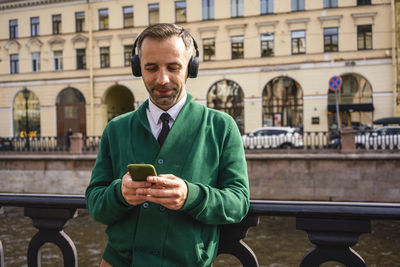 Businessman wearing headphones using mobile phone while standing by railing in city