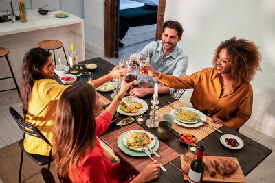 From above side view of cheerful diverse friends celebrating and clinking toasting with wineglasses while sitting at served table with delicious dinner at home