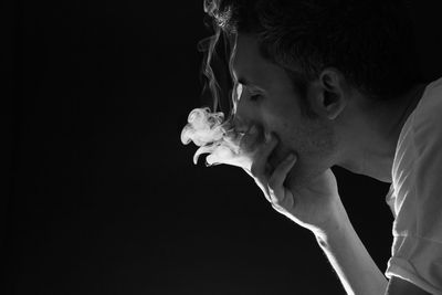 Close-up of man with hand covering mouth emitting smoke against black background