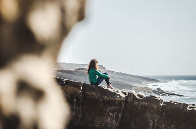 Side view of girl sitting on rocky shore against clear sky