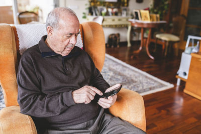 Senior man sitting on armchair at home using smartphone