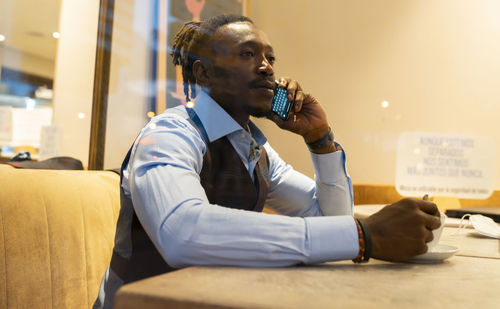 Serious african american male entrepreneur in classy outfit sitting at table with cup of coffee in cafe and speaking on cellphone while looking out of window