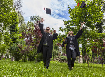 Friends in graduation gown throwing mortarboards against trees