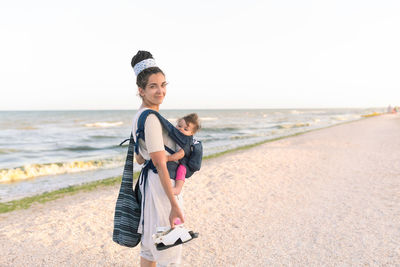 Portrait of mother carrying daughter while standing at beach against sky