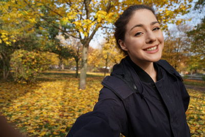 Portrait of smiling young woman standing against trees during autumn