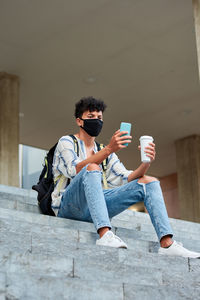 Young afro with mask uses his phone while he is sitting on the stairs