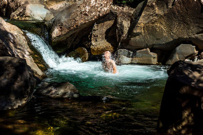 Male tourist emerging from clean pond water near cascade on sunny day in mountains