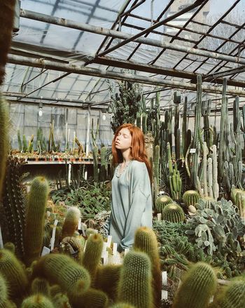 YOUNG WOMAN STANDING AT GREENHOUSE