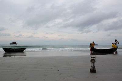 Rear view of people with boat at beach against cloudy sky