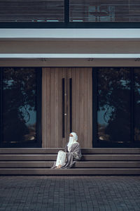 Full length image of veiled woman with white niqab sitting on the stairs in front of the building