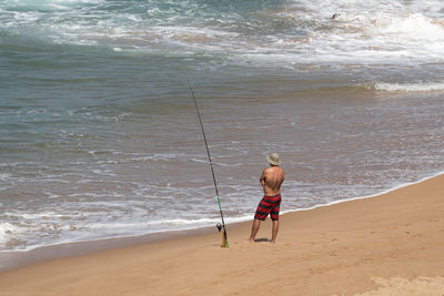 Rear view of shirtless man fishing while standing on shore at beach