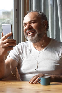 Handsome mature man laughs while has video call with cellphone and headphones at home.