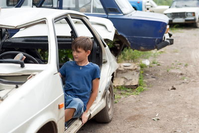 A boy, 11 years old, sits in a disassembled car in a dump of abandoned old cars on a sunny day.