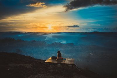 Rear view of woman looking at landscape while sitting on mountain against sky during sunset