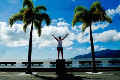 Low angle view of woman with arms raised standing on post amidst palm trees against sea
