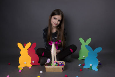 Portrait of smiling girl with eater bunny and pom pom in basket against gray background