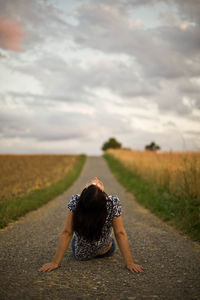 Rear view of woman sitting on road amidst field against cloudy sky