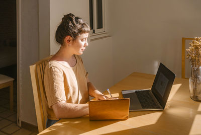 Graphic designer writing in book with laptop at desk