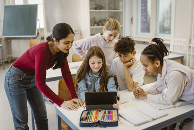 Male and female students doing e-learning with teacher leaning at desk in classroom