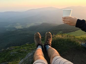 Cropped hand holding disposable cup over legs on mountain