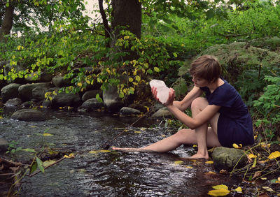 Young woman holding ice while sitting by stream in forest