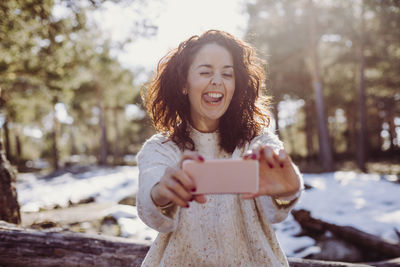 Happy woman taking selfie using mobile phone in forest during winter