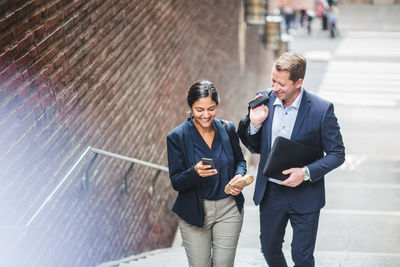 Happy male and female entrepreneurs using phone while climbing staircase in city