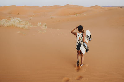 Back view of young female walking on sand and preparing for sandboarding looking away