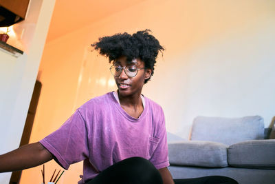 Positive young african american female with afro hair and eyewear modern glasses in casual clothes sitting on floor and painting with brushes while spending time at home during stay at home campaign for coronavirus prevention