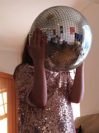 Girl holding disco ball over face against wall at home