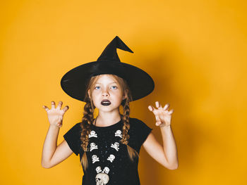 Portrait of girl wearing witch hat gesturing against yellow background during halloween