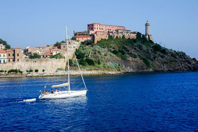 Sailboats sailing on sea by buildings against clear blue sky