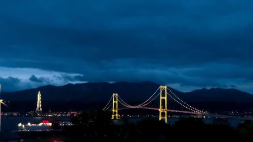 Illuminated suspension bridge against cloudy sky